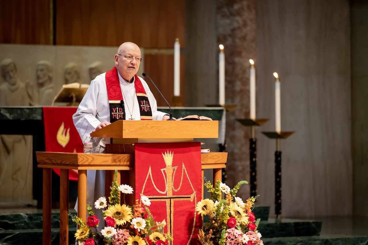 Man in cleric garb preaching at the lecturn inside of the chapel.