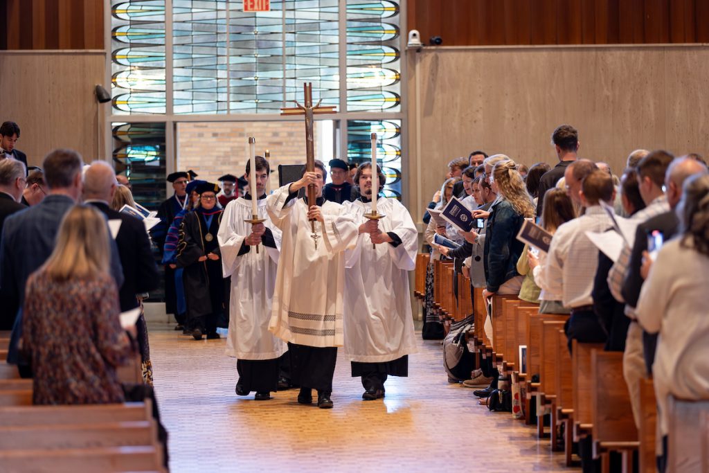 Processional participants in white robes carry a cross down the center aisle of the chapel during Concordia’s Baccalaureate service as attendees stand in the pews.