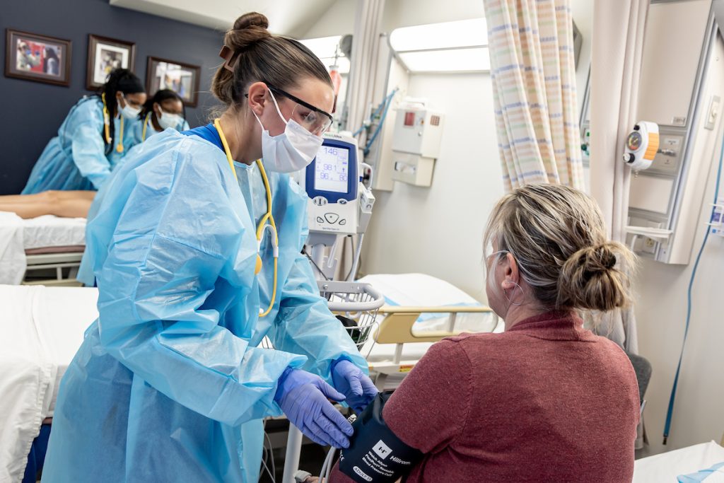 Nursing student in a mask working on a woman who is sitting on a bed.