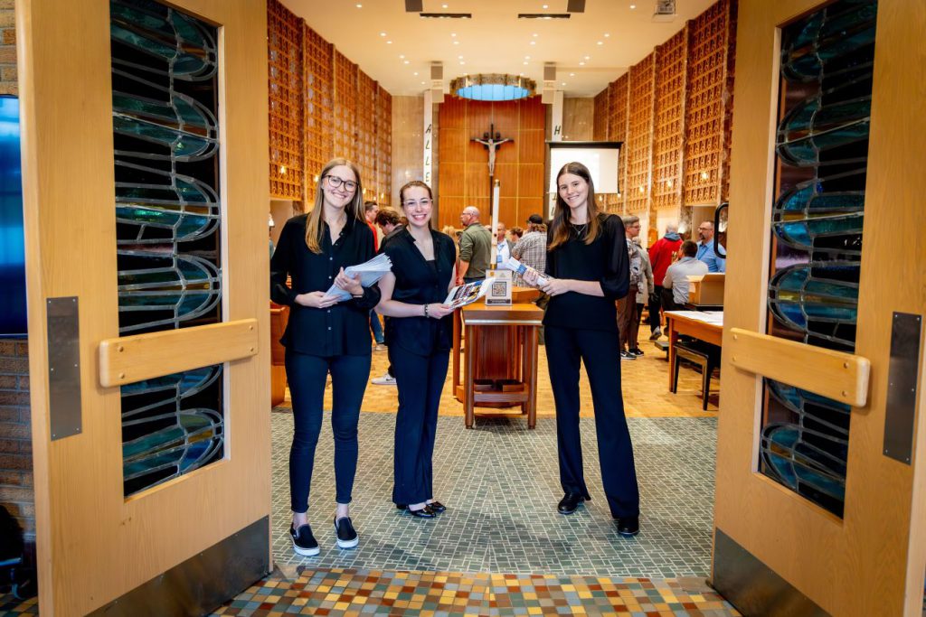 three students standing in front of chapel greeting people