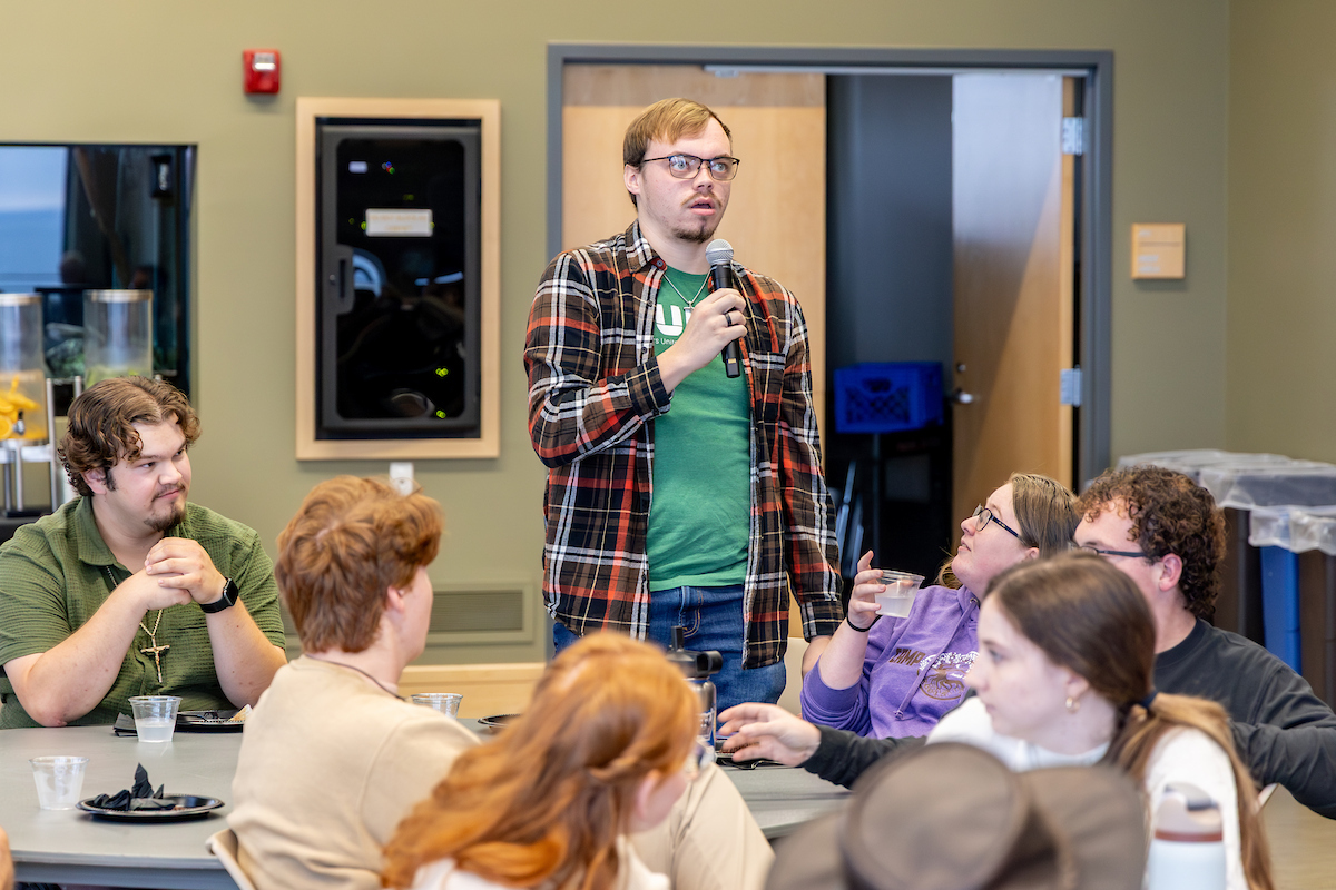 Male student in a green tee with a plaid overshirt stands to talk at a micrphone to students at a circular table.
