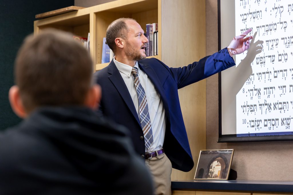 A man in a blue suit points to a screen of foreign language symbols while a student looks on.