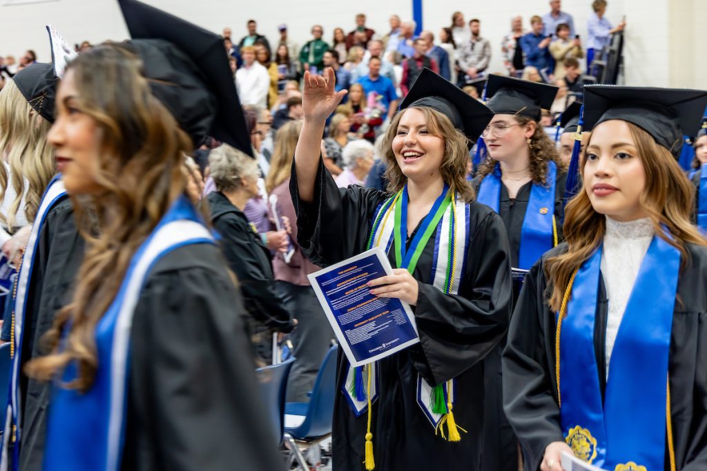 Undergraduate graduates in caps and gowns walk through the ceremony, smiling and holding programs as they process past a seated audience.