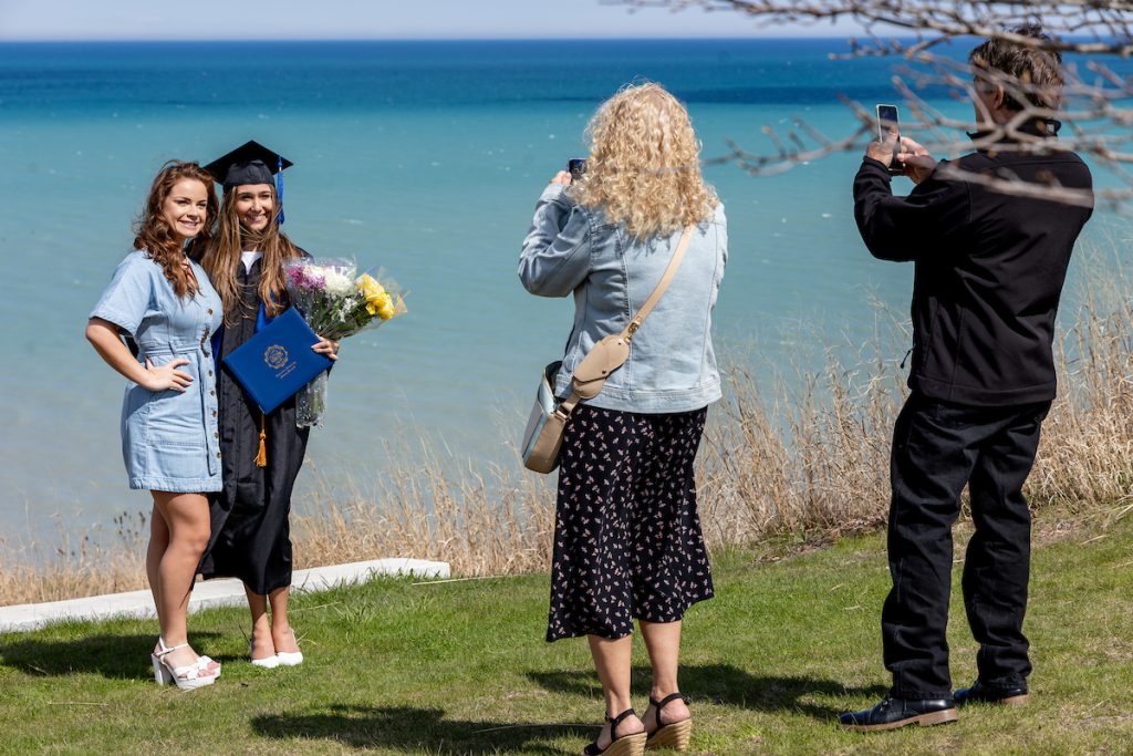Graduate in cap and gown poses for photos with family near Lake Michigan, holding flowers and a diploma cover.
