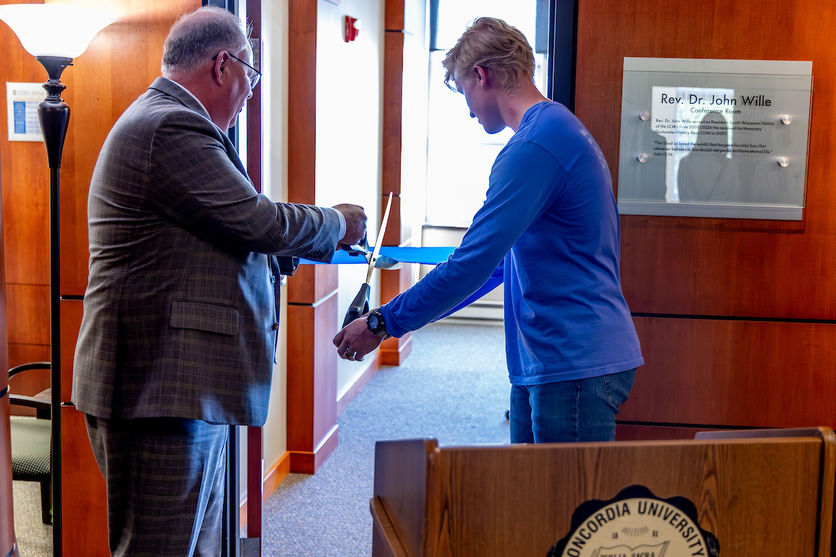 A man in a suit and a man in blue use a large scissors to cut a ribbon across a doorway.